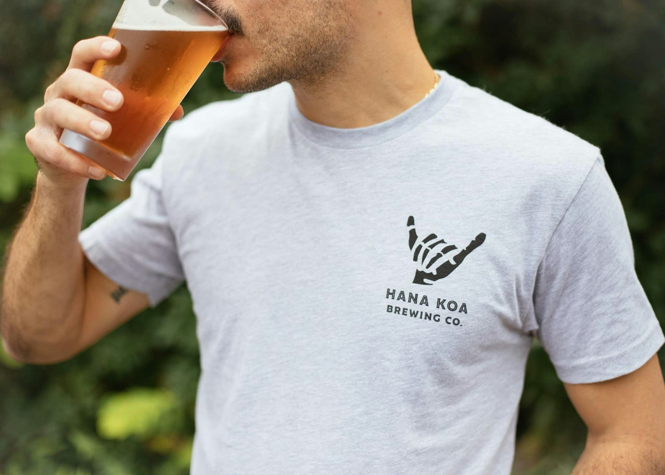 A man takes a sip of beer while wearing a shirt from Hana Koa Brewing