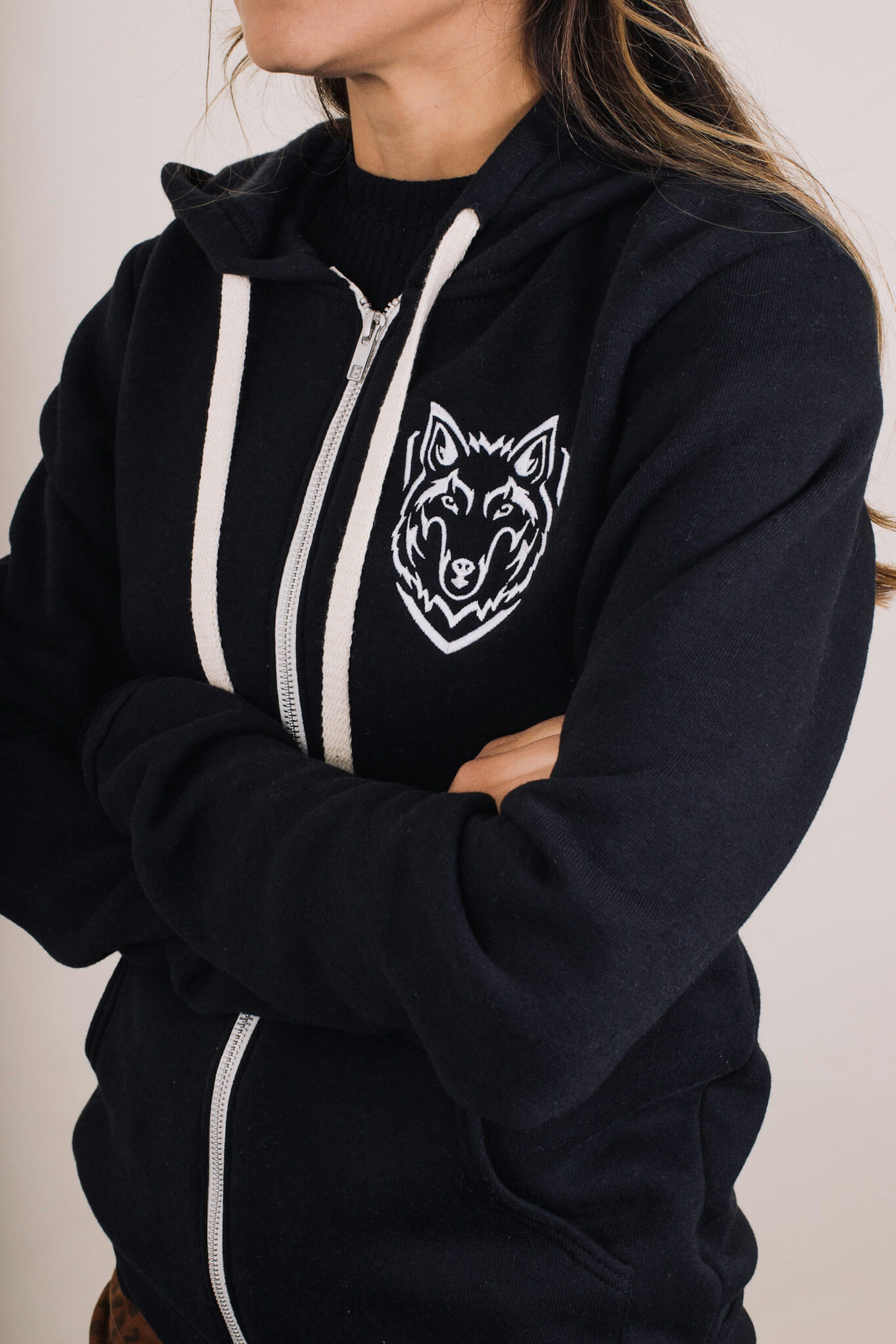 A woman with crossed arms wears a black zippered hoodie with white embroidery on the chest of a wolf