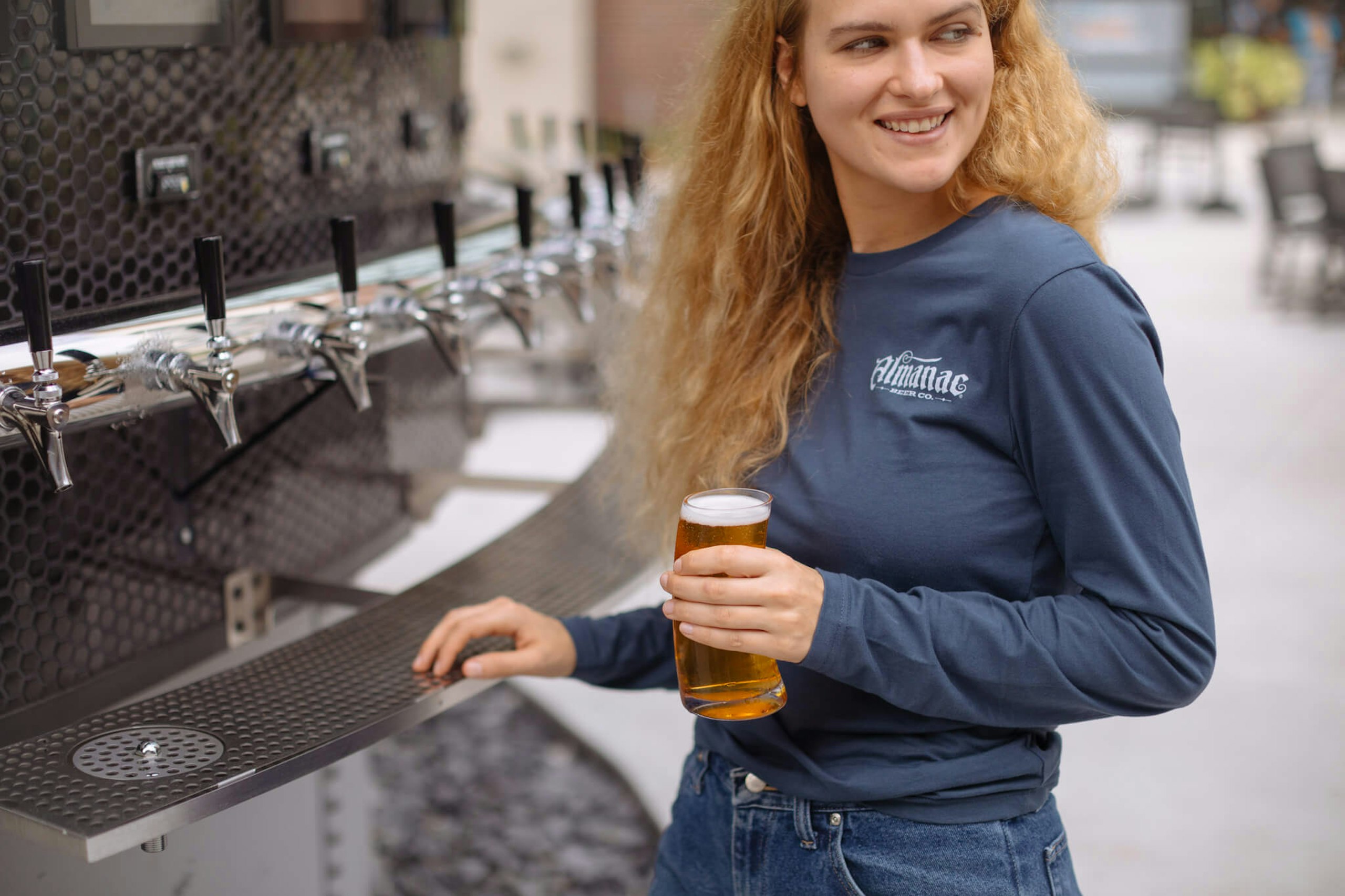 A girl drinks a glass of beer while wearing a long sleeved custom t-shirt with Almanac Beer Co. printed on it