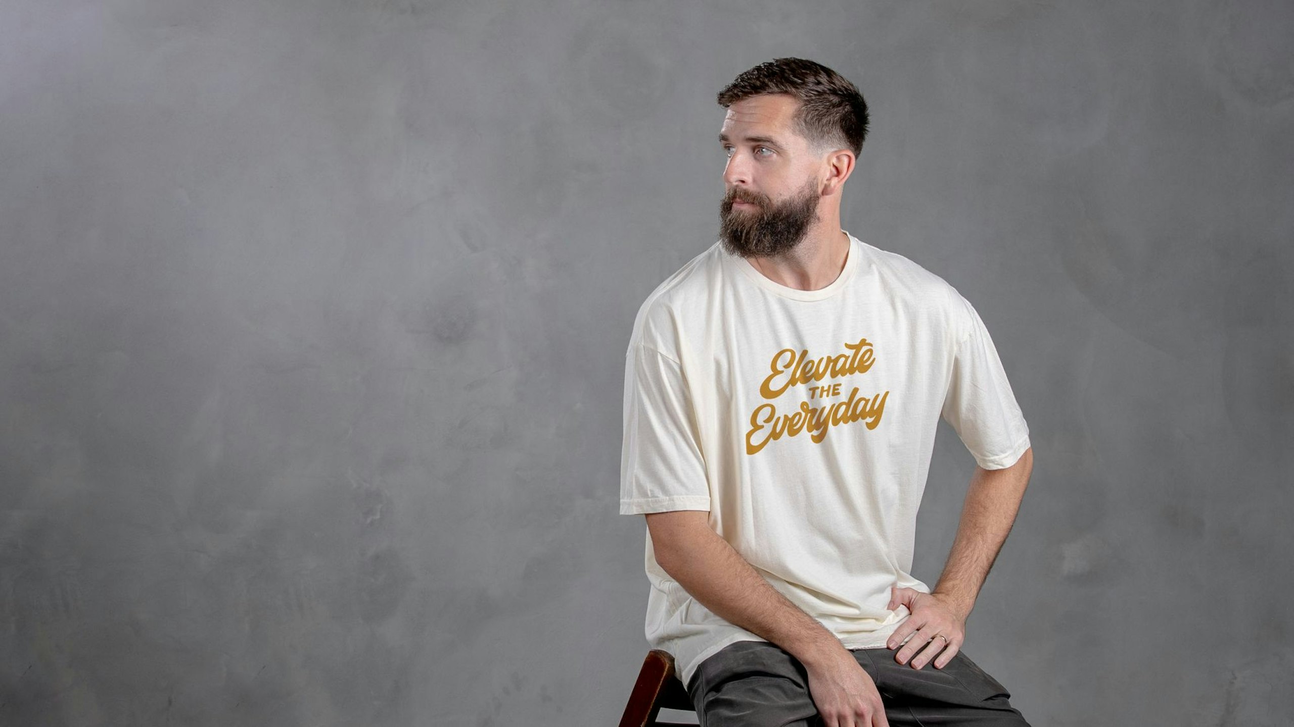 man sitting in front of a dark grey background wearing a cream-white t-shirt with the words elevate the everyday printed on it